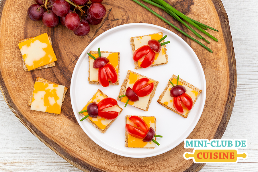 coccinelles faits de craquelins, fromage, tomates pour le corps et raisins pour la tête sur une assiette blanche. Ceci est sur une plaque de bois avec une grappe de raisins et du fromage. 
